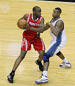 A basketball player, wearing a red jersey with the word «ROCKETS» in the front, is holding the basketball while another basketball player, wearing a white jersey, attempts to steal the ball.